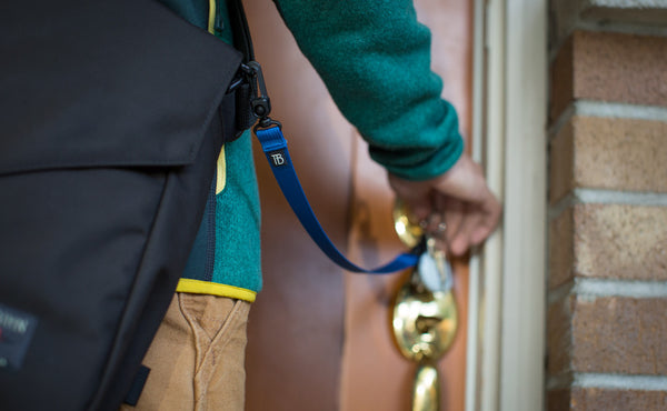A person using a 16-inch Black Key Strap with a snaphook at one end and a split-ring o-ring at the other with a key through the split-ring o-ring to open a door.